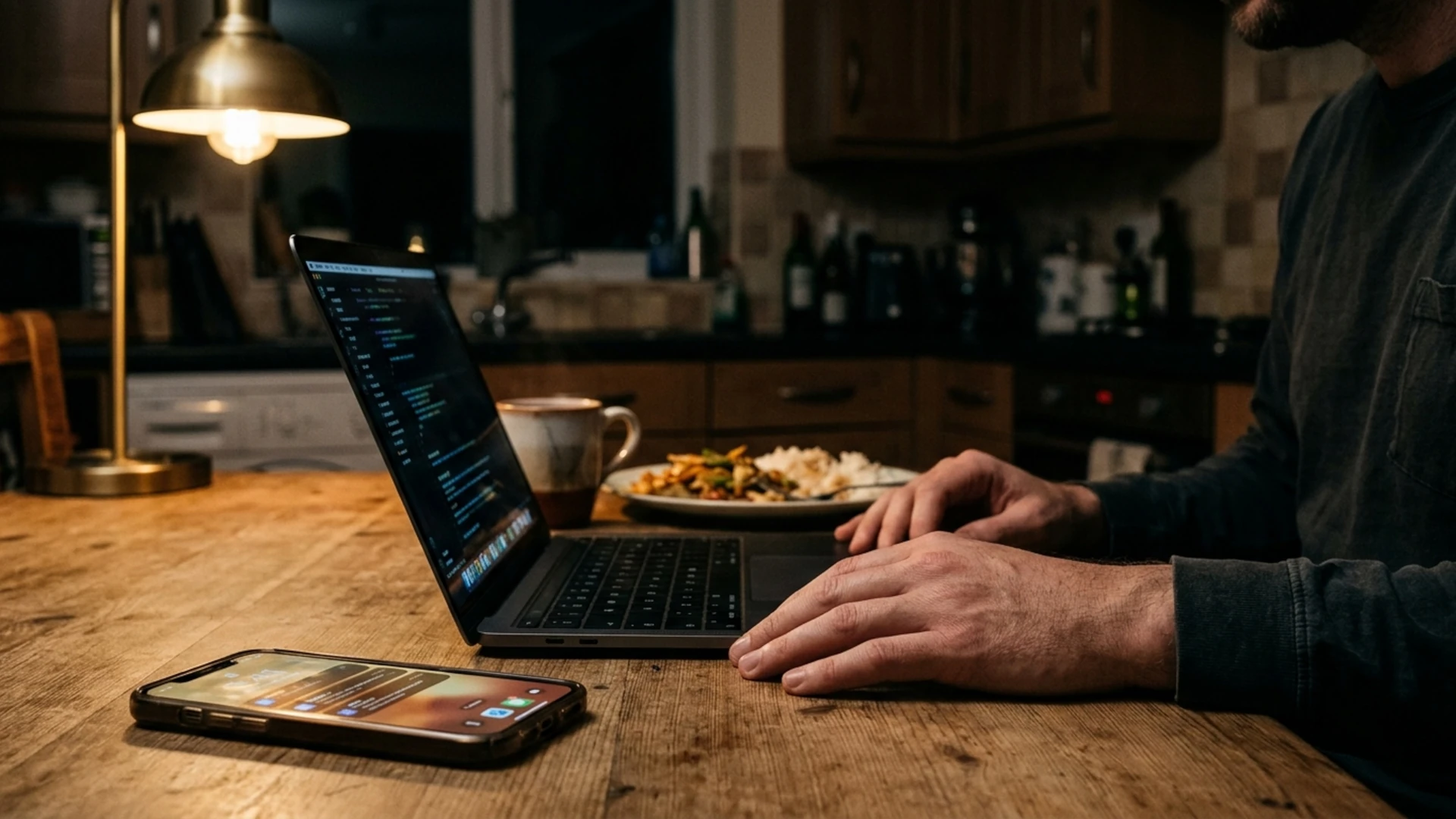 Founder facing the cost of a stalled launch at his kitchen table, phone propped against a laptop, both screens dim, an untouched dinner and mug beside them.