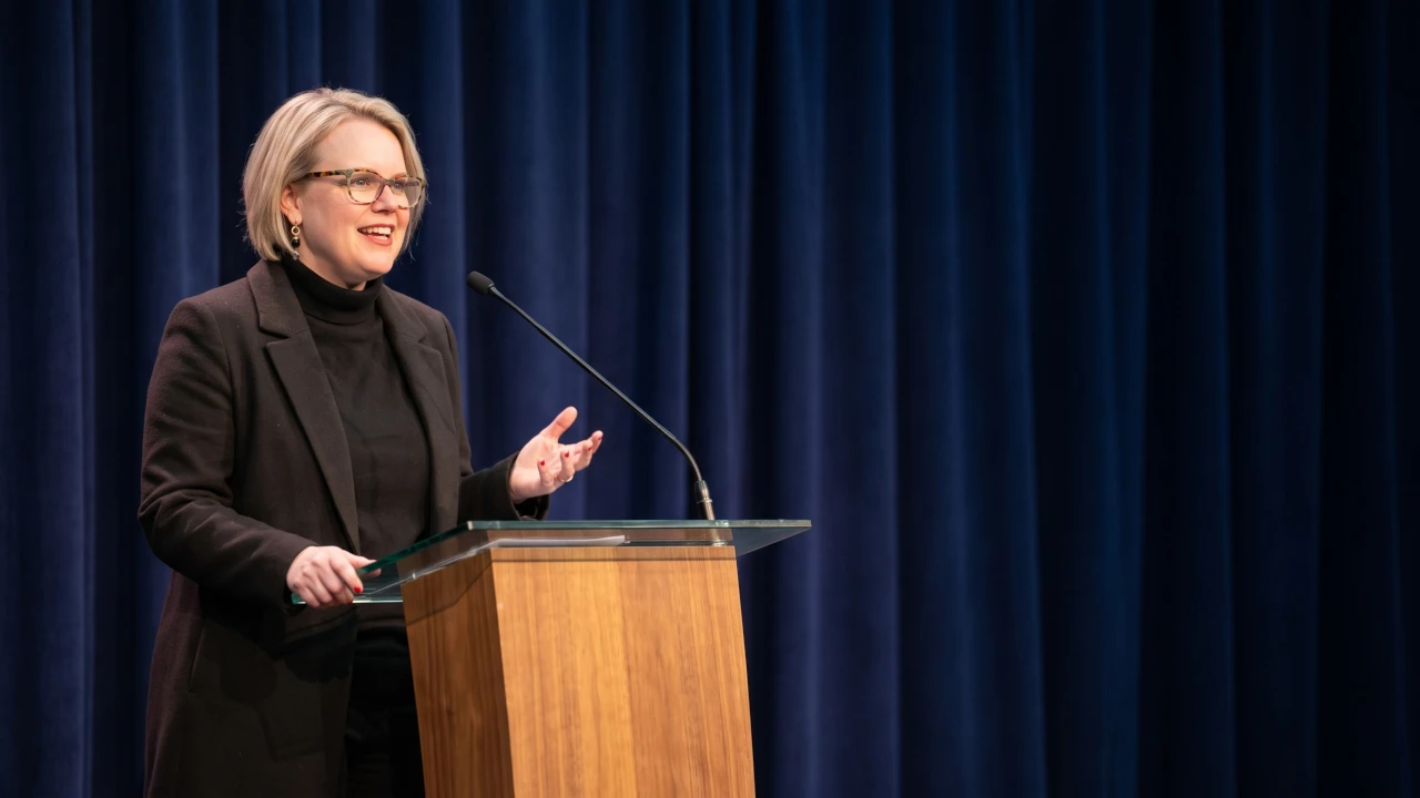 Jualandé Brink mid-sentence behind a wooden lectern, one hand open to land her point, dark blazer over a black turtleneck, deep navy curtain behind her.